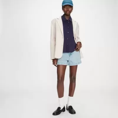 Mulher com camisa azul, blazer bege, calções de ganga, meias brancas, sapatos pretos e boné azul GRACE & MILA