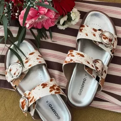 Chinelos Gioseppo com padrão castanho e branco e fivelas prateadas ao lado de flores