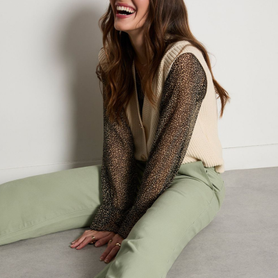 Mulher sentada vestindo blusa preta transparente com padrão de bolinhas brancas, colete creme de malha e calças verdes claras.