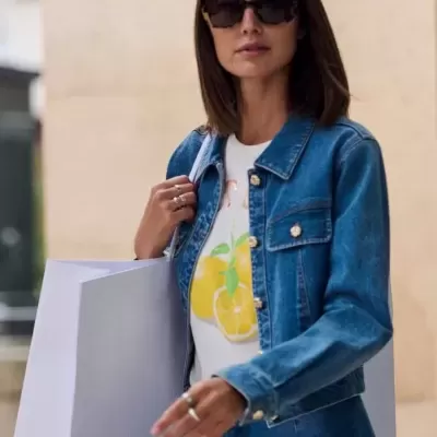 Mulher com casaco e calças de ganga azuis, t-shirt branca com limões, óculos de sol escuros e sacola branca