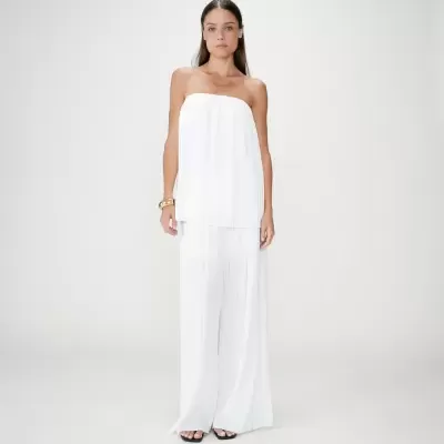 Mulher com conjunto branco pregueado e sandálias pretas em fundo cinza claro.