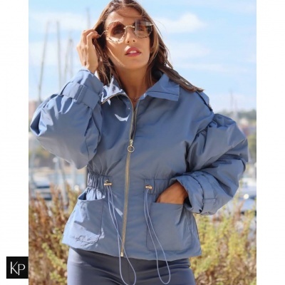 Casaco azul céu com fecho de correr e bolsos visto numa mulher junto a um porto com barcos