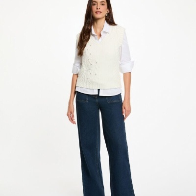 Mulher a usar camisa branca, colete branco de malha, calças de ganga azuis e sapatos pretos num fundo branco