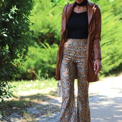 Mulher com casaco castanho, calças de padrão pele de cobra e chapéu bege a caminhar numa zona exterior com vegetação.