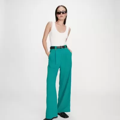 Mulher com blusa branca, calças verdes largas, cinto e sapatos pretos, óculos de sol, fundo branco