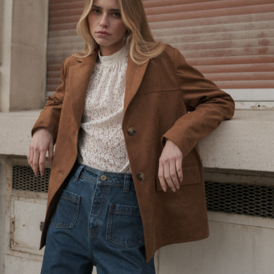 Mulher com casaco castanho, blusa branca de renda e calças de ganga azuis encostada num muro de betão