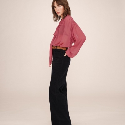 Mulher com blusa rosa transparente e calças pretas com cinto castanho num fundo neutro