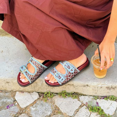 Sandálias femininas trançadas azul/castanho com fivelas prateadas e vestido castanho sentada em degrau com copo de bebida.