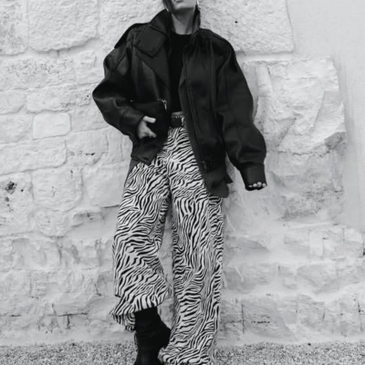 Mulher com casaco preto oversize, calças com padrão zebra e botas pretas junto a muro branco.