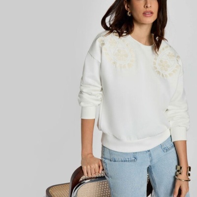 Mulher com sweatshirt branca bordada e jeans azuis claros encostada numa cadeira de metal com assento em palhinha