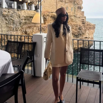 Mulher com roupa bege, chapéu estampado e bolsa bege numa varanda com vista para o mar