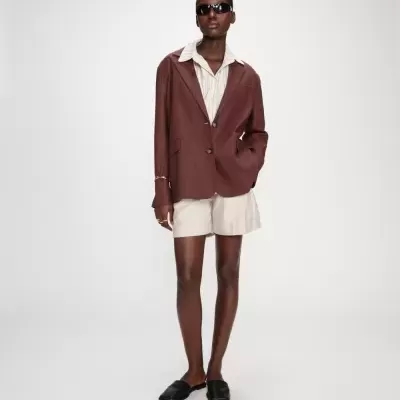 Pessoa com blazer castanho, camisa creme, calções creme e sapatos mule pretos com óculos de sol.
