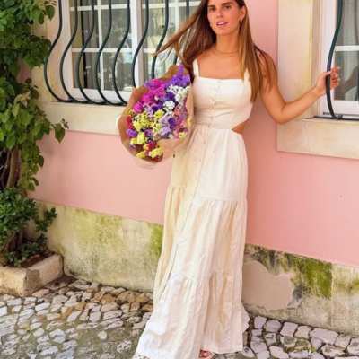 Mulher com vestido comprido branco e sandálias brancas segurando ramo de flores coloridas em fundo de parede rosa com janela