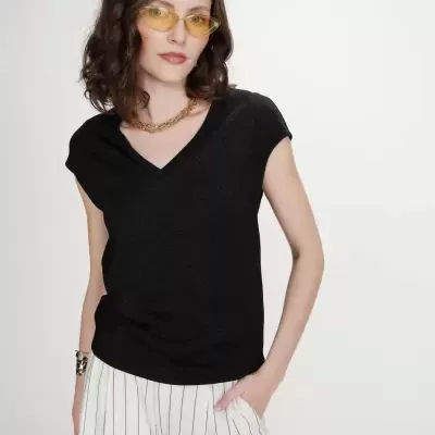 Mulher com camisola preta de gola em V e calças brancas às riscas, óculos de sol amarelos e colar dourado.