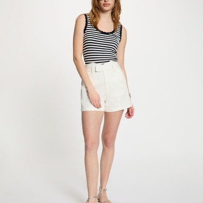 Mulher de pé com top às riscas preto e branco, calções brancos, sandálias prateadas e óculos de sol pretos sobre fundo branco.