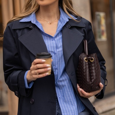 Mulher com trench coat azul, camisa riscada azul, jeans, óculos de sol pretos, boné bege e bolsa castanha de couro trançado segurando café.