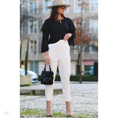 Mulher com blusa preta, calças brancas, chapéu bege, óculos e mala preta numa rua urbana