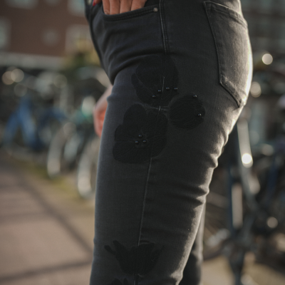 Calças pretas de ganga com padrão floral em relevo e botões, mãos no bolso, cenário urbano com bicicletas