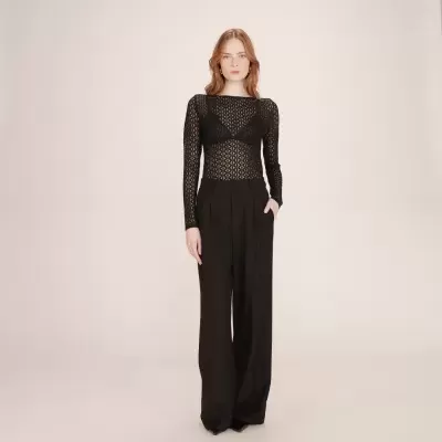 Mulher vestida com top preto transparente e calças pretas largas em fundo neutro.