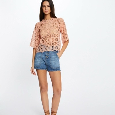 Mulher veste camisa rosa claro em renda floral, calções de ganga azuis e sandálias castanhas, fundo branco