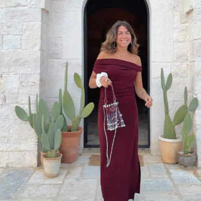 Mulher com vestido longo bordô e mala transparente com franjas em frente a entrada de pedra com cactos