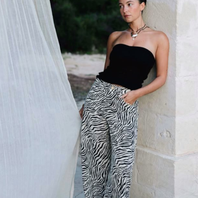 Mulher com top preto e calças de padrão zebra encostada em parede de pedra clara com cortina branca ao lado