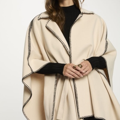 Capa bege com costuras pretas em ziguezague, camisola preta e calças de pele.