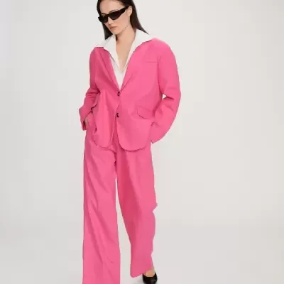 Mulher vestindo blazer e calças cor-de-rosa, camisa branca, sapatos pretos, óculos escuros, fundo branco