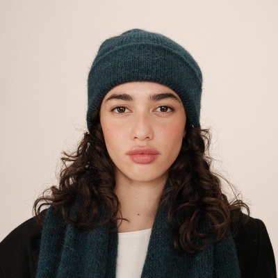 Mulher com gorro e echarpe azul-petróleo, casaco preto e fundo bege