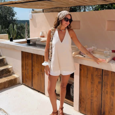 Mulher com roupa de verão branca e sandálias castanhas numa cozinha exterior com armários de madeira.