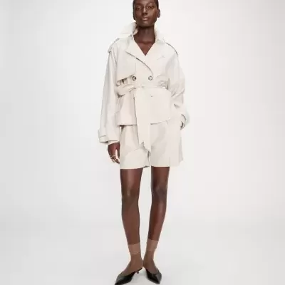 Mulher vestindo conjunto bege claro composto por blazer com botões e cinto e bermuda, com meias castanhas e sapatos pretos.