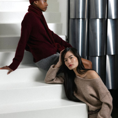 Duas mulheres com roupas de malha grosso bordô e castanho claro sentadas numa escada branca, fundo com paredes metálicas.