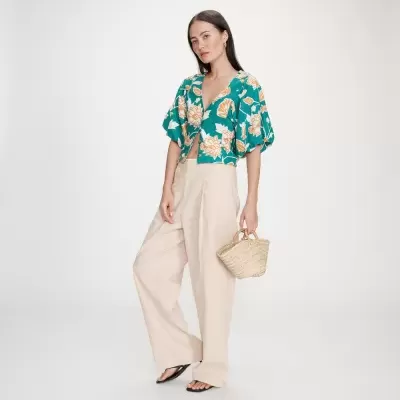 Mulher com blusa floral verde, calças bege largas, sandálias pretas e bolsa de palha pequena.