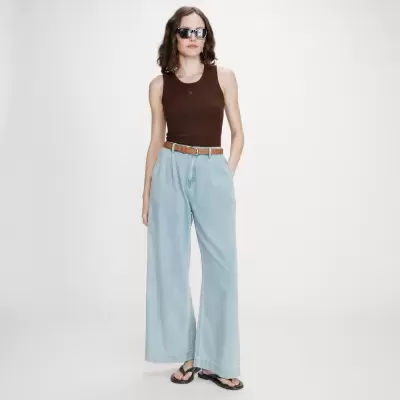Mulher com top castanho, calças de ganga largas claras, cinto castanho e chinelos pretos.