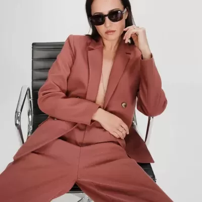 Mulher com fato castanho avermelhado e óculos sentada em cadeira de escritório preta.