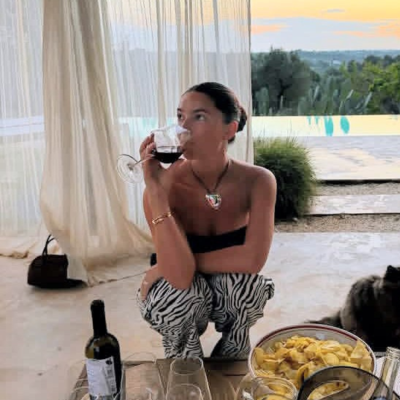 Mulher a beber vinho tinto junto a mesa com bebidas e snacks, ao ar livre com cortina branca e piscina ao fundo