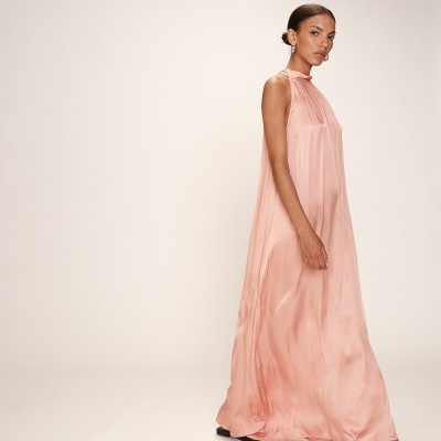 Vestido comprido cor-de-rosa fluído de gola alta e sem mangas com sapatos pretos, mulher de cabelo apanhado.