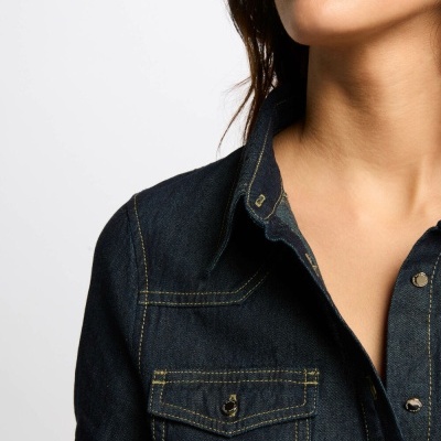 Camisa de ganga azul escura com costuras amarelas e bolso de botão, usada por mulher