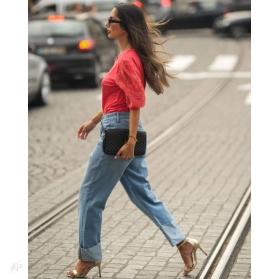 Mulher veste blusa rosa, calças de ganga e sandálias douradas enquanto caminha numa rua com calçada de pedras e carris.