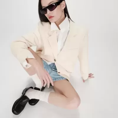 Mulher sentada com blazer bege, saia de ganga, meias brancas, sapatos pretos e óculos de sol.