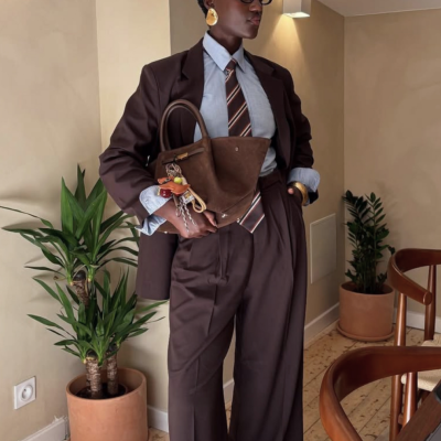 Mulher com blazer e calças castanho escuro, camisa azul, gravata riscada, mala castanha, plantas em vasos e cadeira de madeira ao lado.