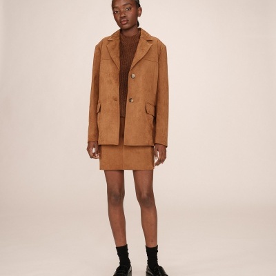 Mulher vestindo casaco e saia castanhos, meias e sapatos pretos, em fundo neutro