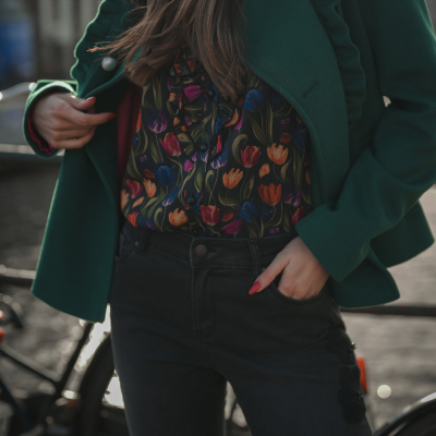 Mulher a usar casaco verde, camiseta floral e calças pretas com bicicleta ao fundo