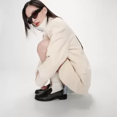 Mulher agachada com blazer bege, saia jeans, meias brancas, mocassins pretos e óculos de sol pretos num fundo branco.