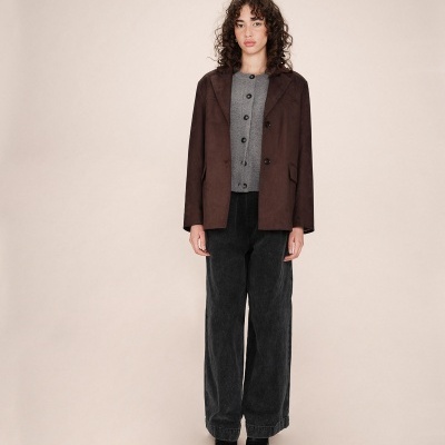 Mulher com casaco castanho, blusa cinzenta e calças pretas largas em fundo bege claro