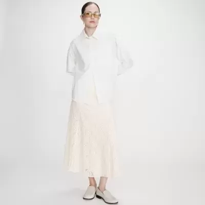 Mulher com camisa branca, saia midi creme em renda, sapatos bége claros e óculos de sol amarelos.