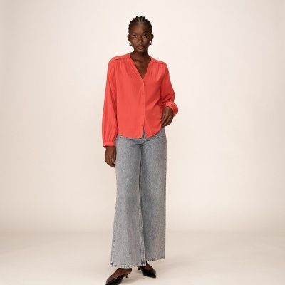 Mulher com blusa coral, calças de ganga claras e sapatos pretos em fundo neutro