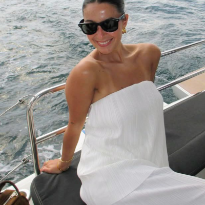 Mulher de vestido branco alças, óculos escuros e acessórios dourados sentado em banco de barco com mar ao fundo