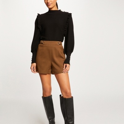 Mulher a usar camisola preta com folhos e calções castanhos com botas pretas altas.