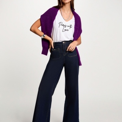 Mulher com calças largas azul escuras, t-shirt branca com texto, cardigan roxo e botas pretas em fundo branco